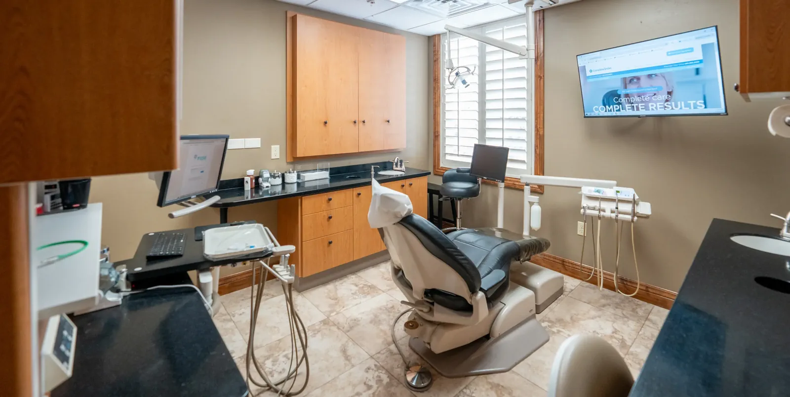 Dental exam room with a patient chair, medical equipment, wooden cabinets, and a wall-mounted television.